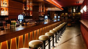 Un bar con una barra larga, taburetes y estantes llenos de botellas.