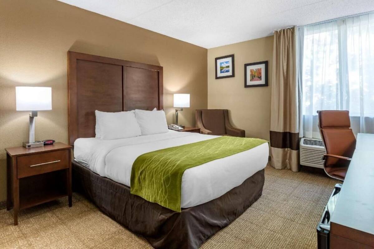 A hotel room with a bed, two bedside lamps, a chair, a desk, and a window with curtains.