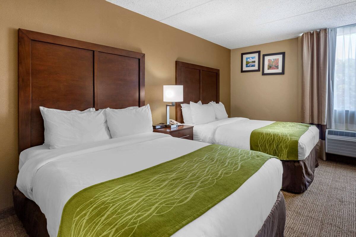A hotel room with two beds, a nightstand, a lamp, and a window with curtains.