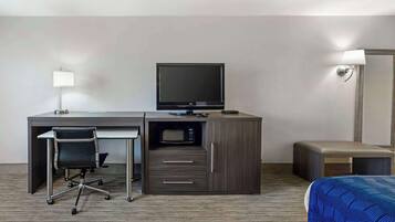Habitación de hotel con escritorio, silla, televisor, microondas, cama con colcha azul estampada y mesita de noche.