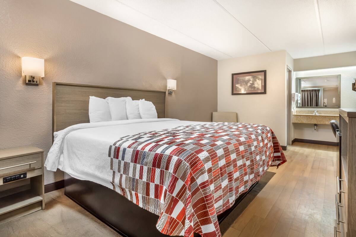 A hotel room with a large bed, bedside tables, a nightstand, a bathroom with a mirror, and a wall-mounted light fixture.