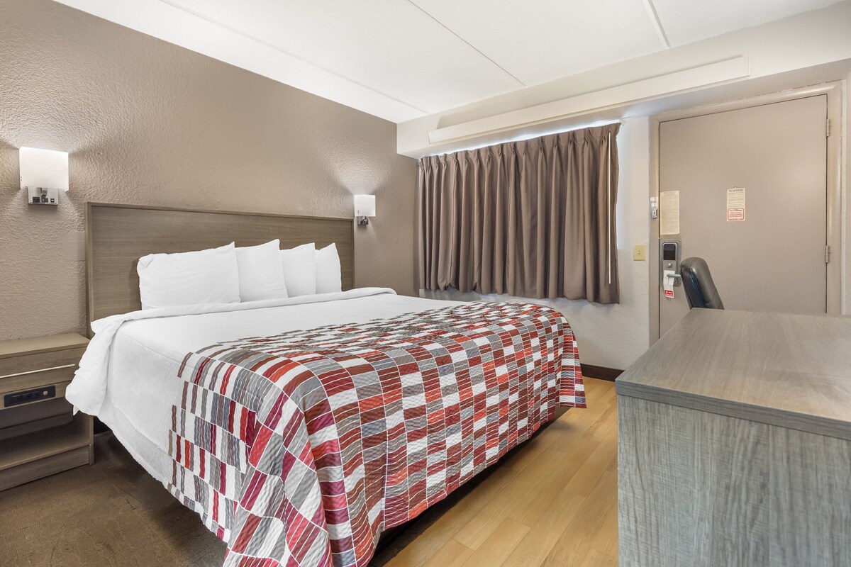 A hotel room with a large bed, a desk, and a chair. There is a wall-mounted light and a window with curtains.