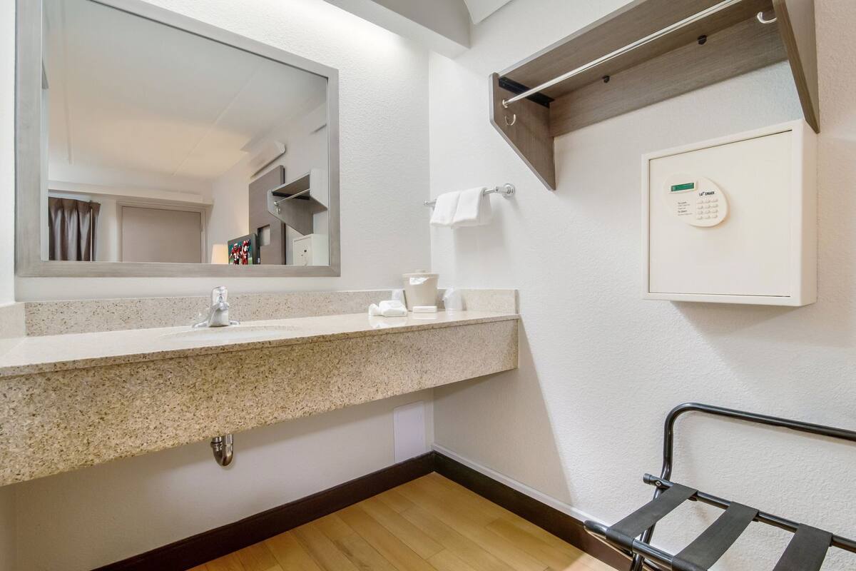 A bathroom with a granite countertop sink, a mirror, a towel rack, a towel, a soap dispenser, and a wall-mounted thermostat.