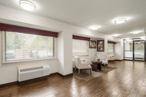 A spacious hallway with wooden flooring, white walls, and multiple windows with blinds. There are two white chairs and a small table with a basket.