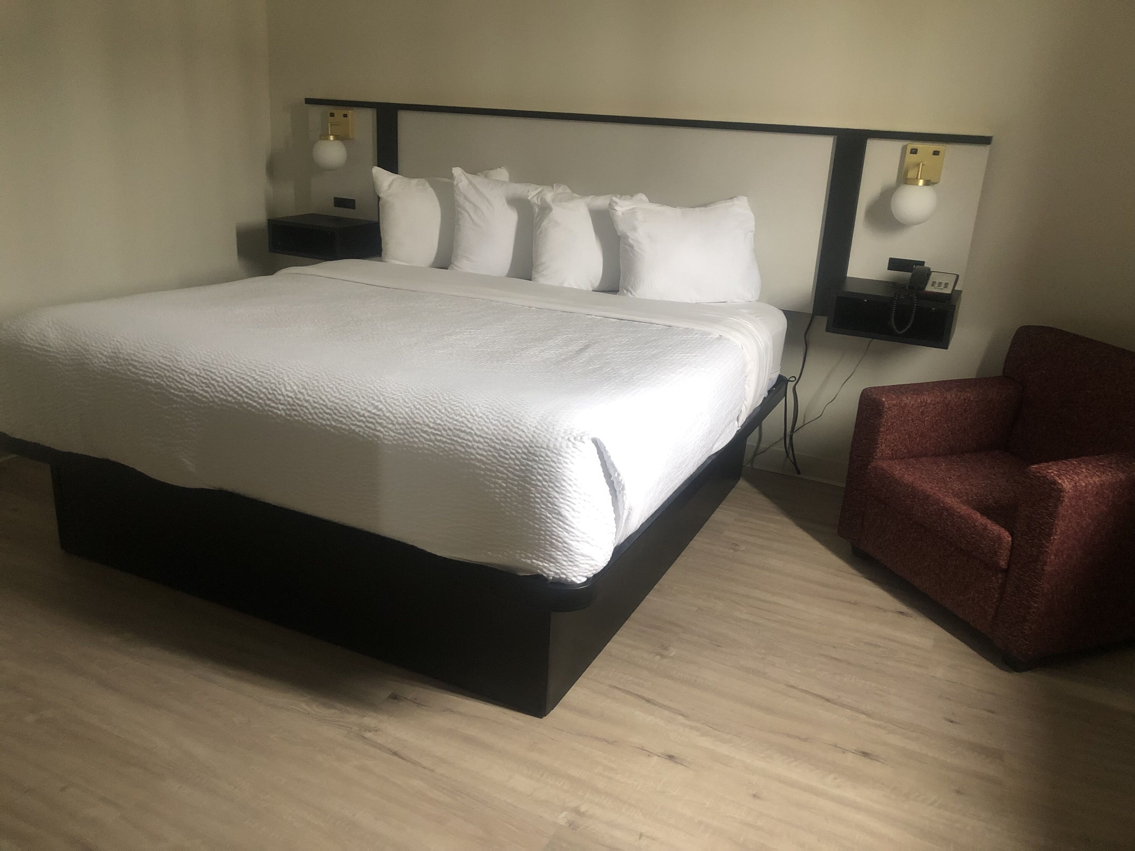 Ein ordentlich bezogenes Bett mit weißen Laken, einem dunklen Kopfteil und einem roten Sessel zur rechten Seite.
