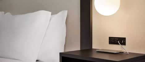 A neatly made bed with white linens, a bedside table with a smartphone and charger, and a wall-mounted light fixture.