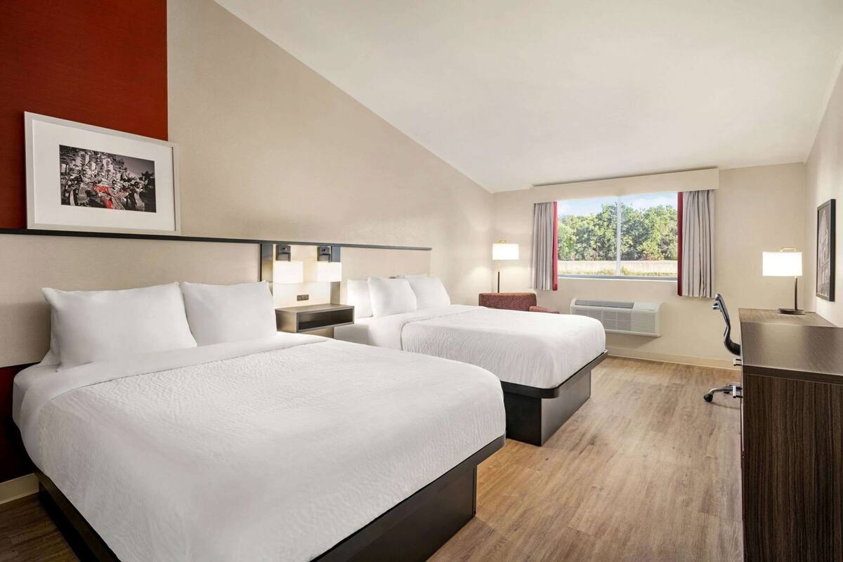 Een hotelkamer met twee bedden, een bureau, een stoel, een raam met gordijnen en een ingelijste foto aan de muur.