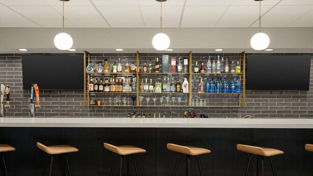 A bar area with a black brick wall, shelves of liquor bottles, and four stools. - Ramada by Wyndham Allentown/Bethlehem (Whitehall)