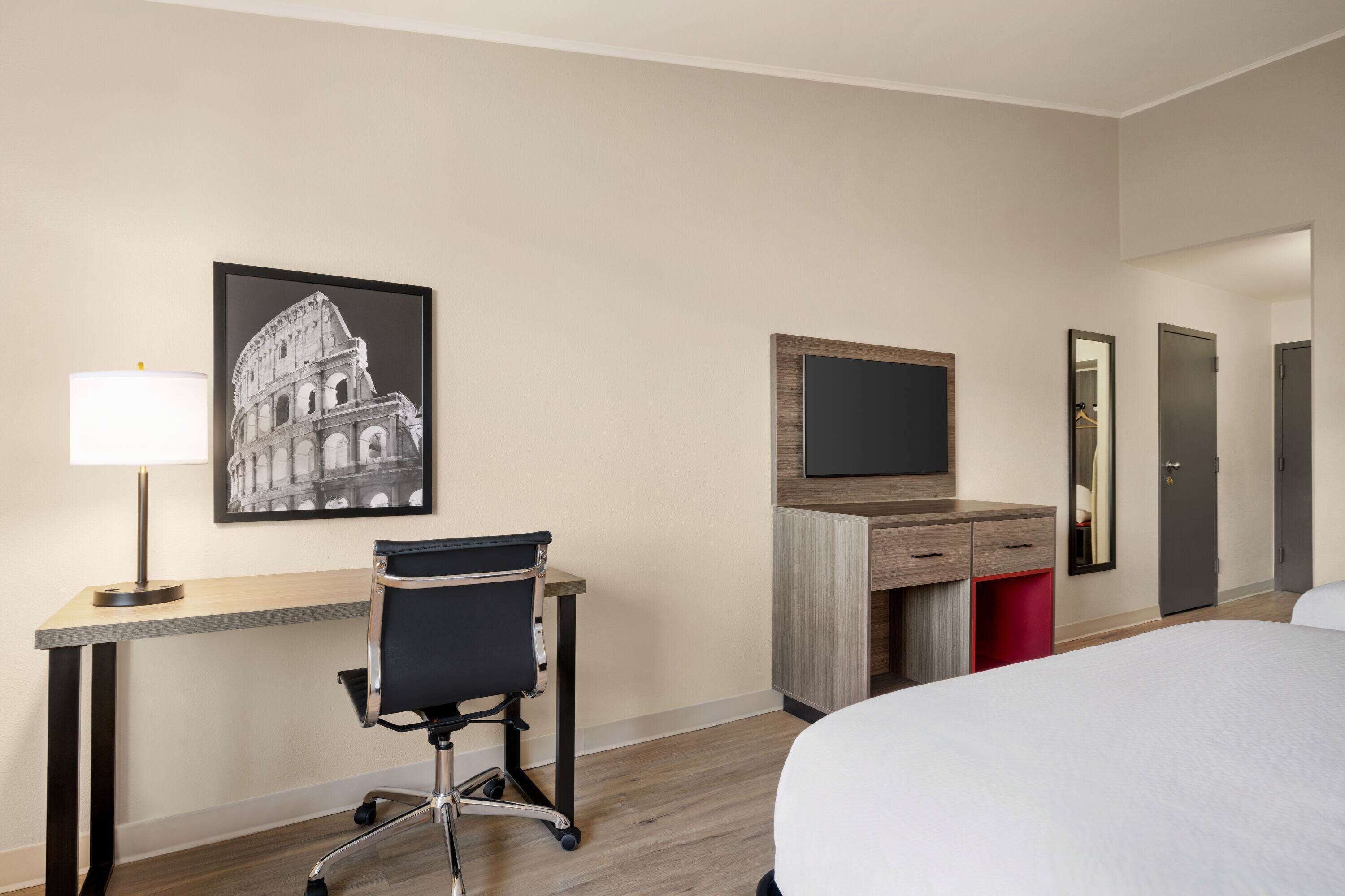 Ett hotellrum med en säng, ett skrivbord, en stol, en TV, en garderob och en inramad tavla som föreställer Colosseum.