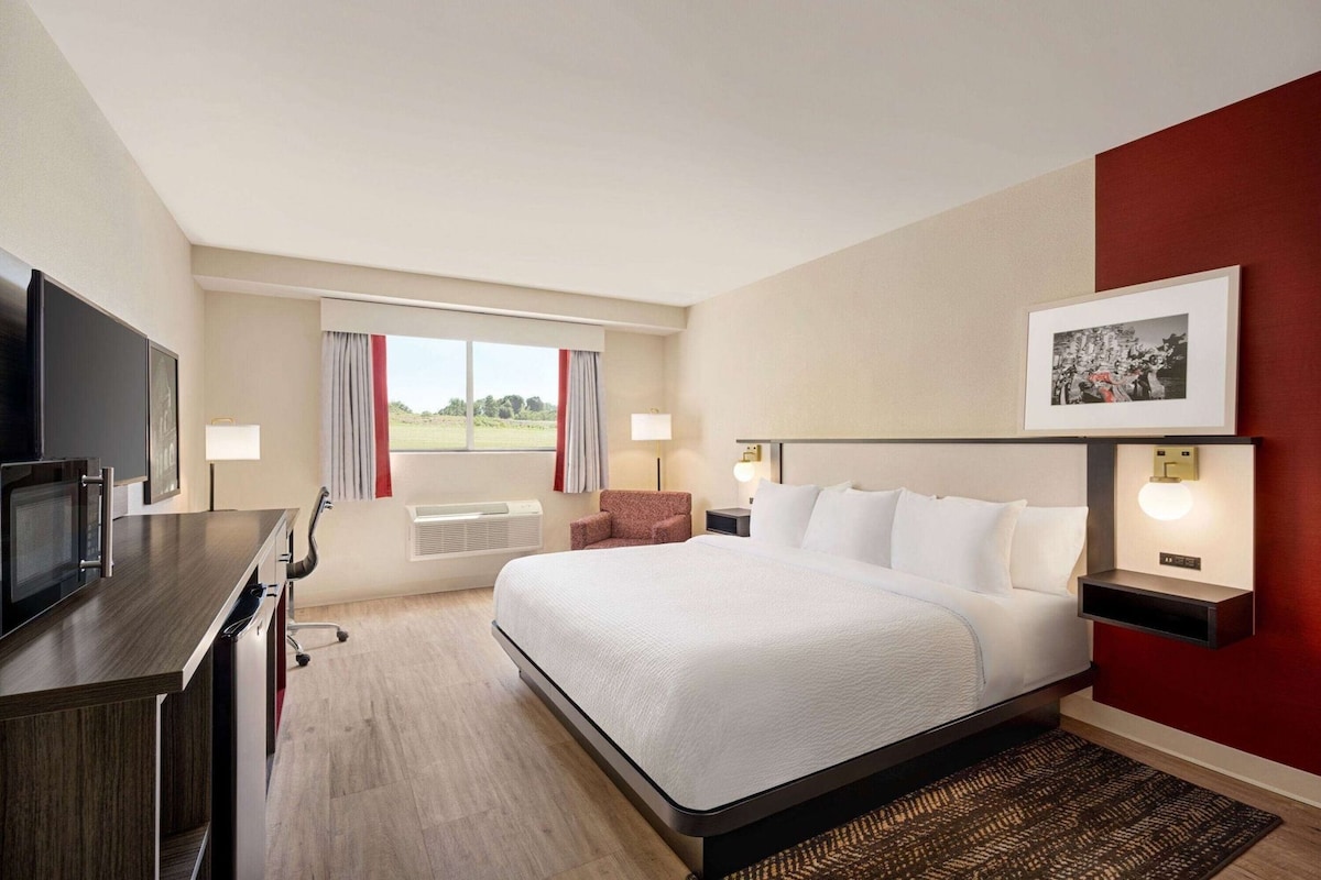 Een hotelkamer met een bed, een bureau, een televisie, een raam met gordijnen en een ingelijst schilderij aan de muur.