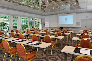Ein Konferenzraum mit mehreren Tischen und Stühlen, einer Projektionsleinwand, die einen Hotelnamen zeigt, und großen Fenstern mit Blick auf Grünflächen.