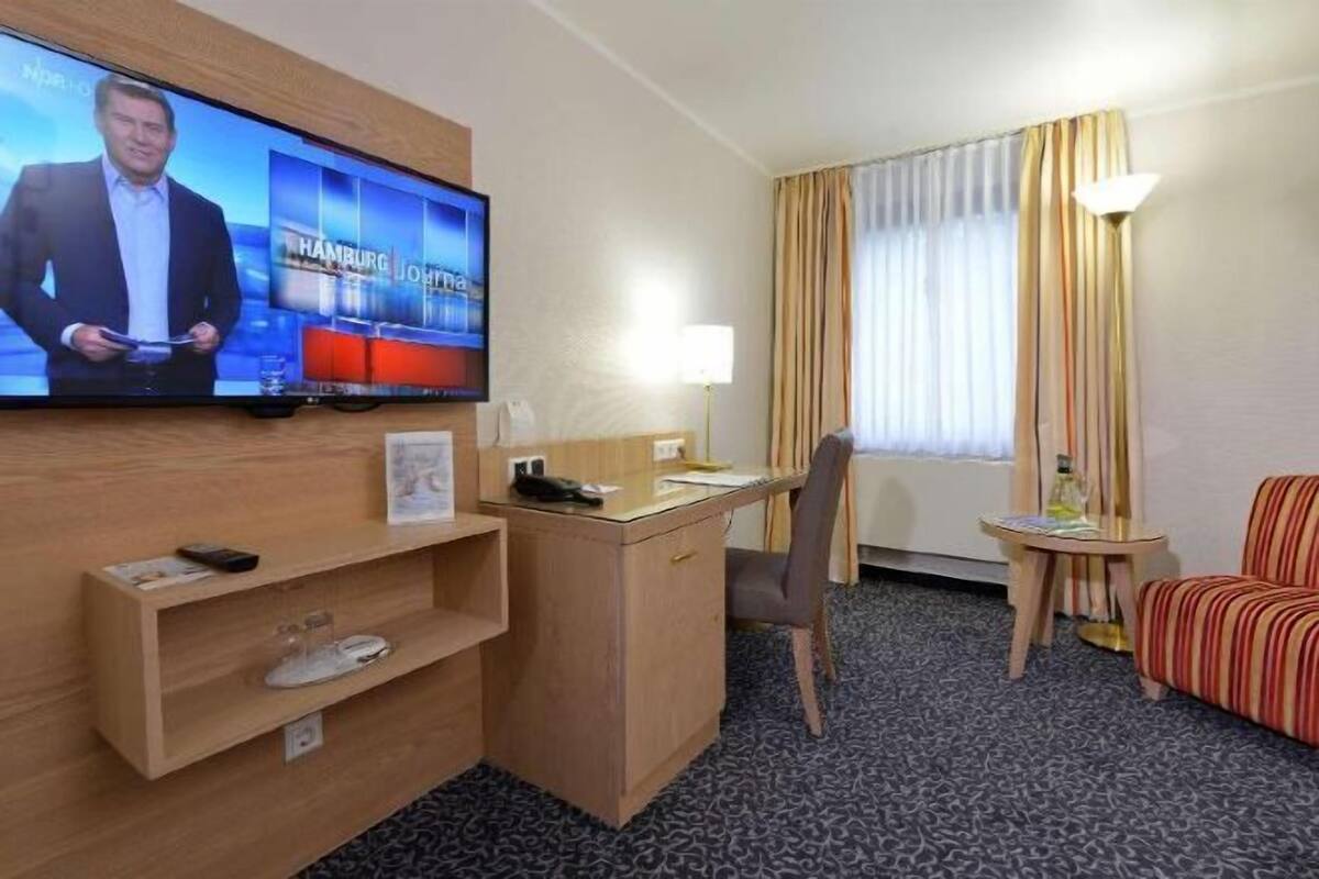 Une chambre d’hôtel équipée d’une télévision diffusant une chaîne d’information, d’un bureau avec une lampe, d’une chaise, d’une petite table et d’un canapé.