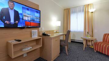 A hotel room with a television displaying a news program, a desk with a telephone, a chair, a small table with a lamp, and a striped armchair.
