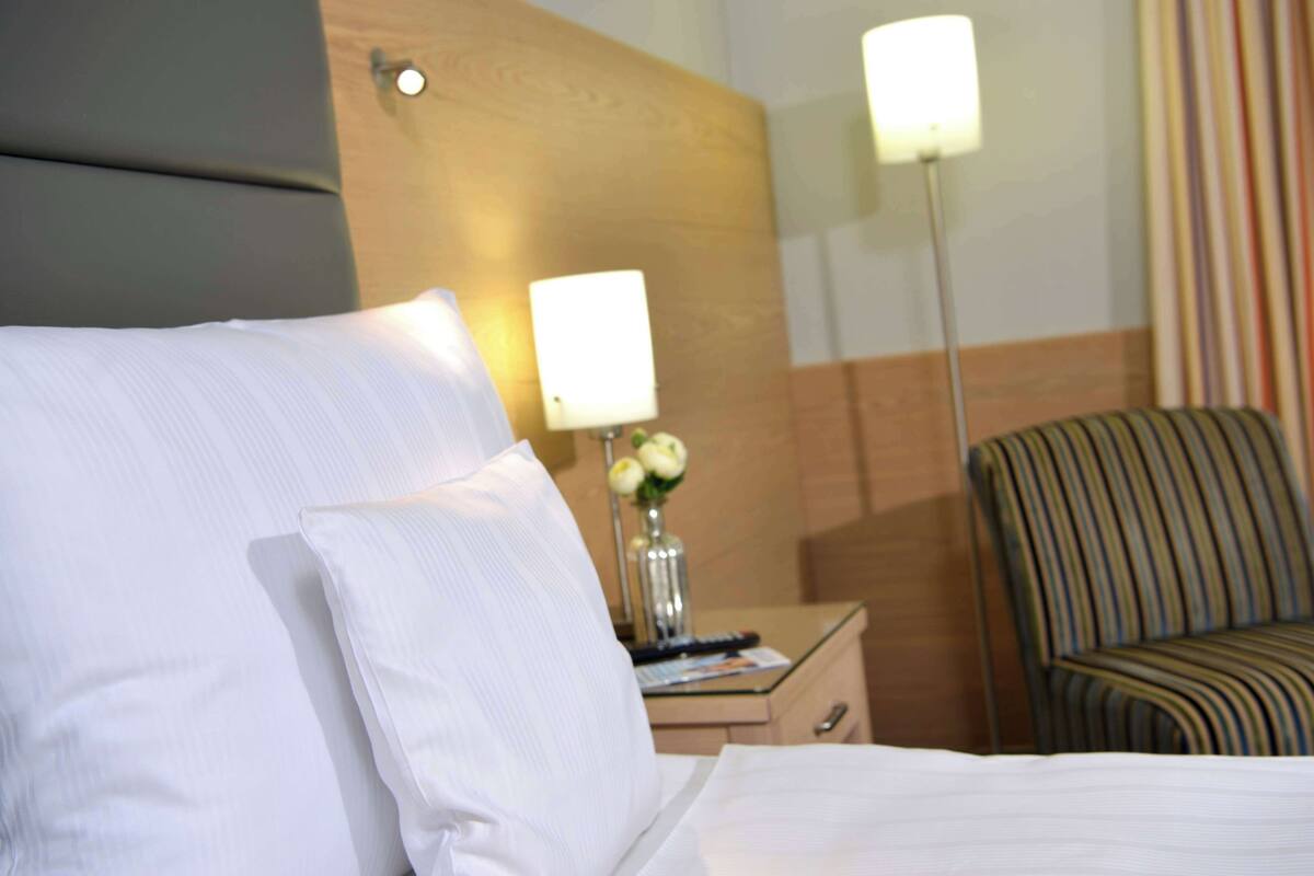 Une chambre d’hôtel avec un lit, une table de chevet avec une lampe, un vase de fleurs et une chaise à rayures.