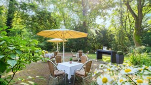 Une terrasse extérieure avec une table et des chaises, entourée d’une végétation luxuriante et d’arbres.