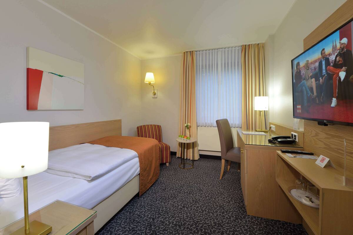 Une chambre d’hôtel avec un lit, un bureau, une chaise, une télévision et une petite table avec une lampe.