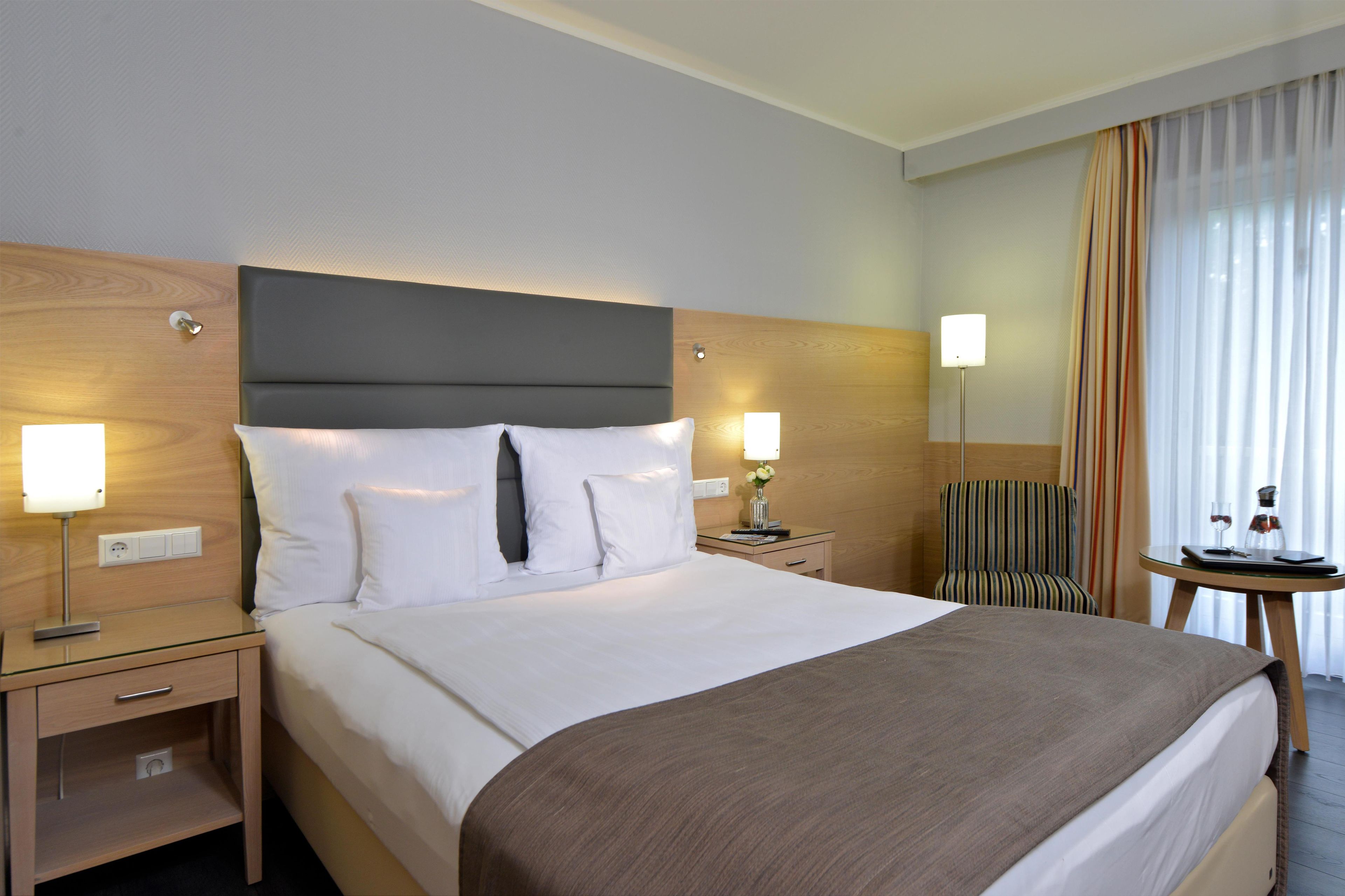 Ein Hotelzimmer mit einem großen Bett, Nachttischen, einem Sessel, einem kleinen Tisch mit Lampe und einem Fenster mit Vorhängen.
