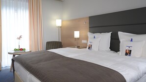 Une chambre d’hôtel avec un grand lit, une table de chevet avec une lampe, une chaise et une petite table sur laquelle se trouvent des fruits et un vase.