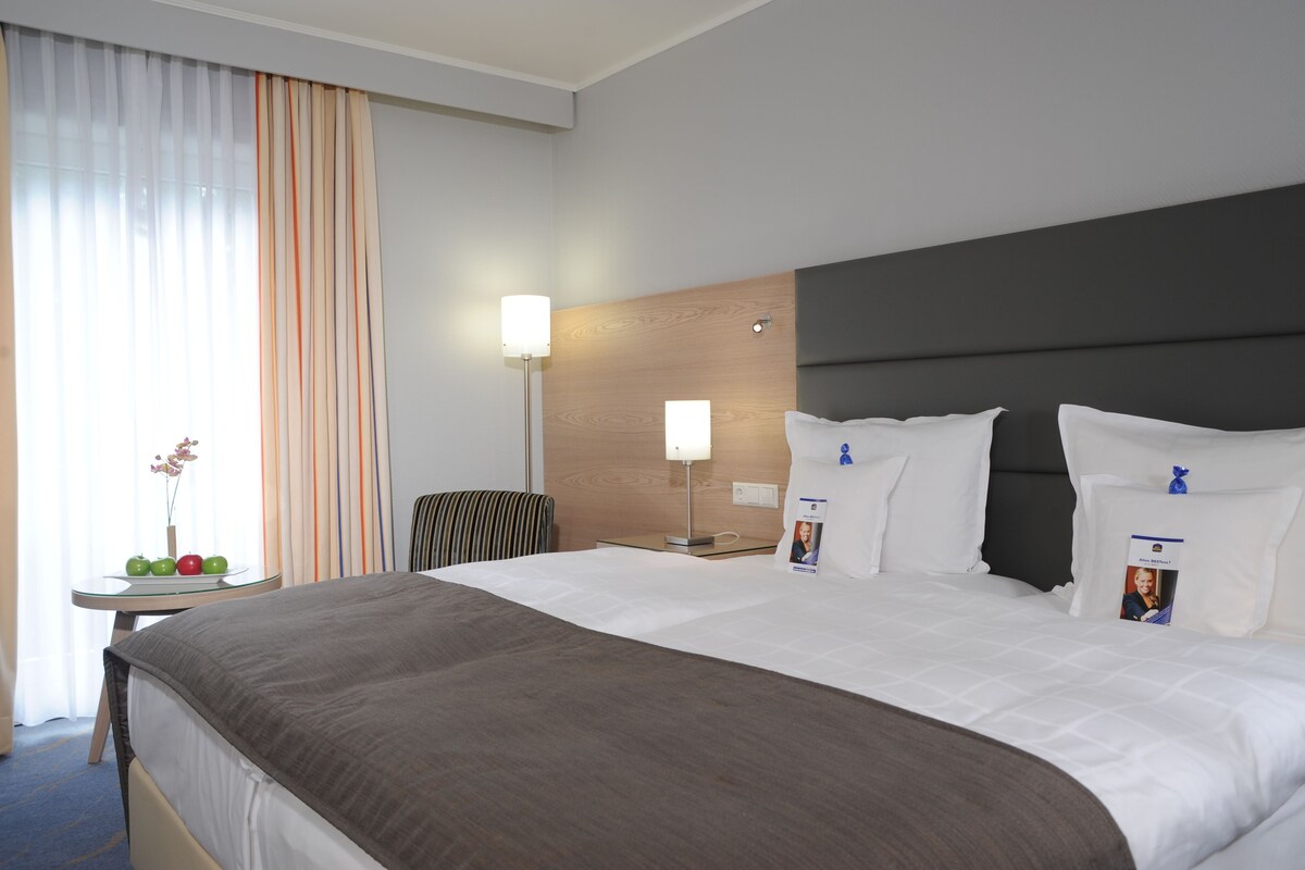 A hotel room with a large bed, a bedside table with a lamp, a chair, and a small table with fruit and a vase.