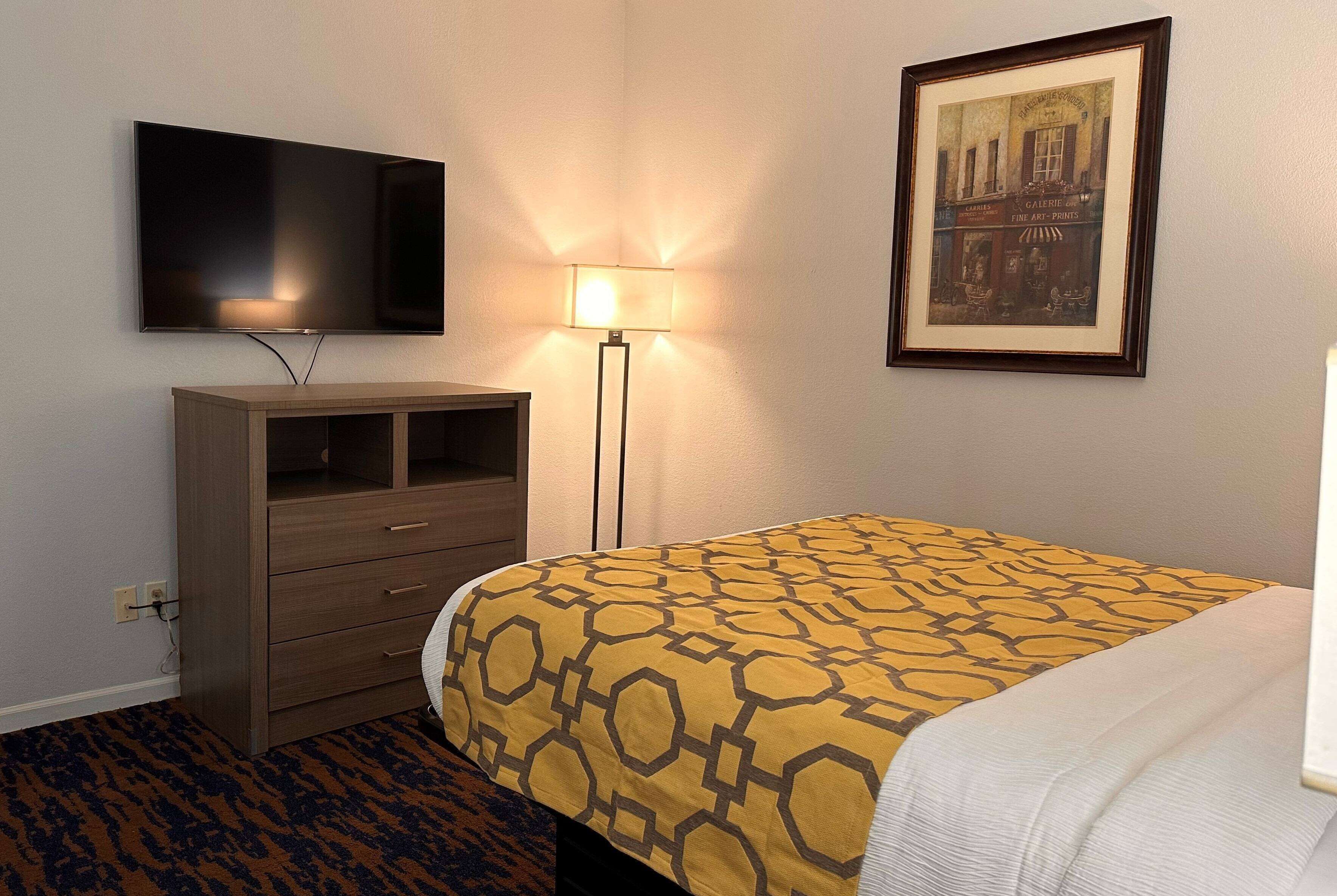 A hotel room with a bed, a television mounted on the wall, a desk with drawers, a lamp, and a framed picture on the wall.