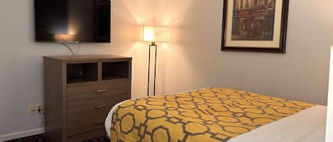 A hotel room with a bed, a television mounted on the wall, a desk with drawers, a lamp, and a framed picture on the wall.