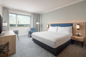 A hotel room with a large bed, a desk with a chair, a small table, and a view of the cityscape. - Hilton Paris Charles de Gaulle Airport (Tremblay-en-France)