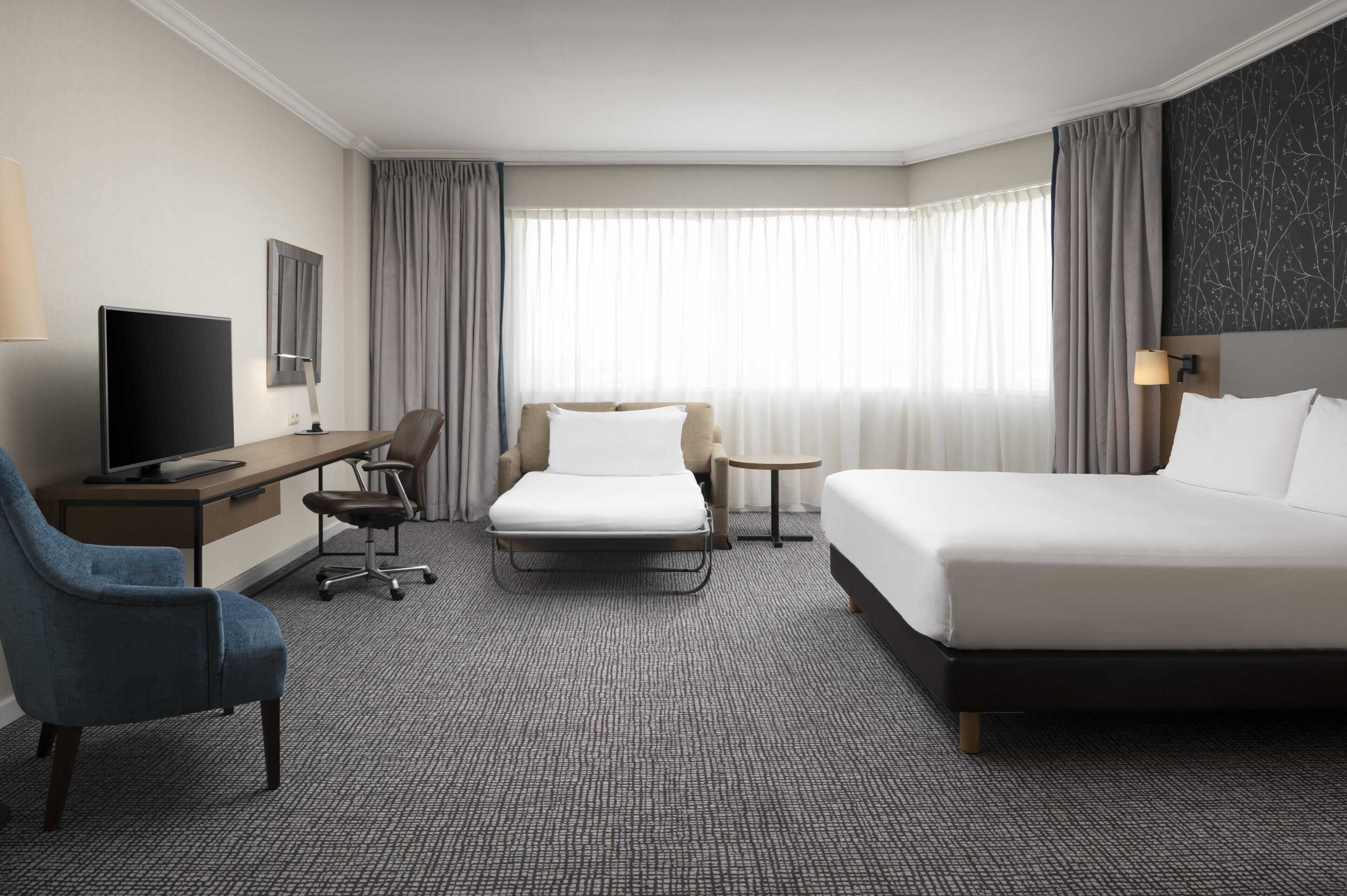 A hotel room with a bed, a desk with a TV, a chair, a small table, and a sofa.