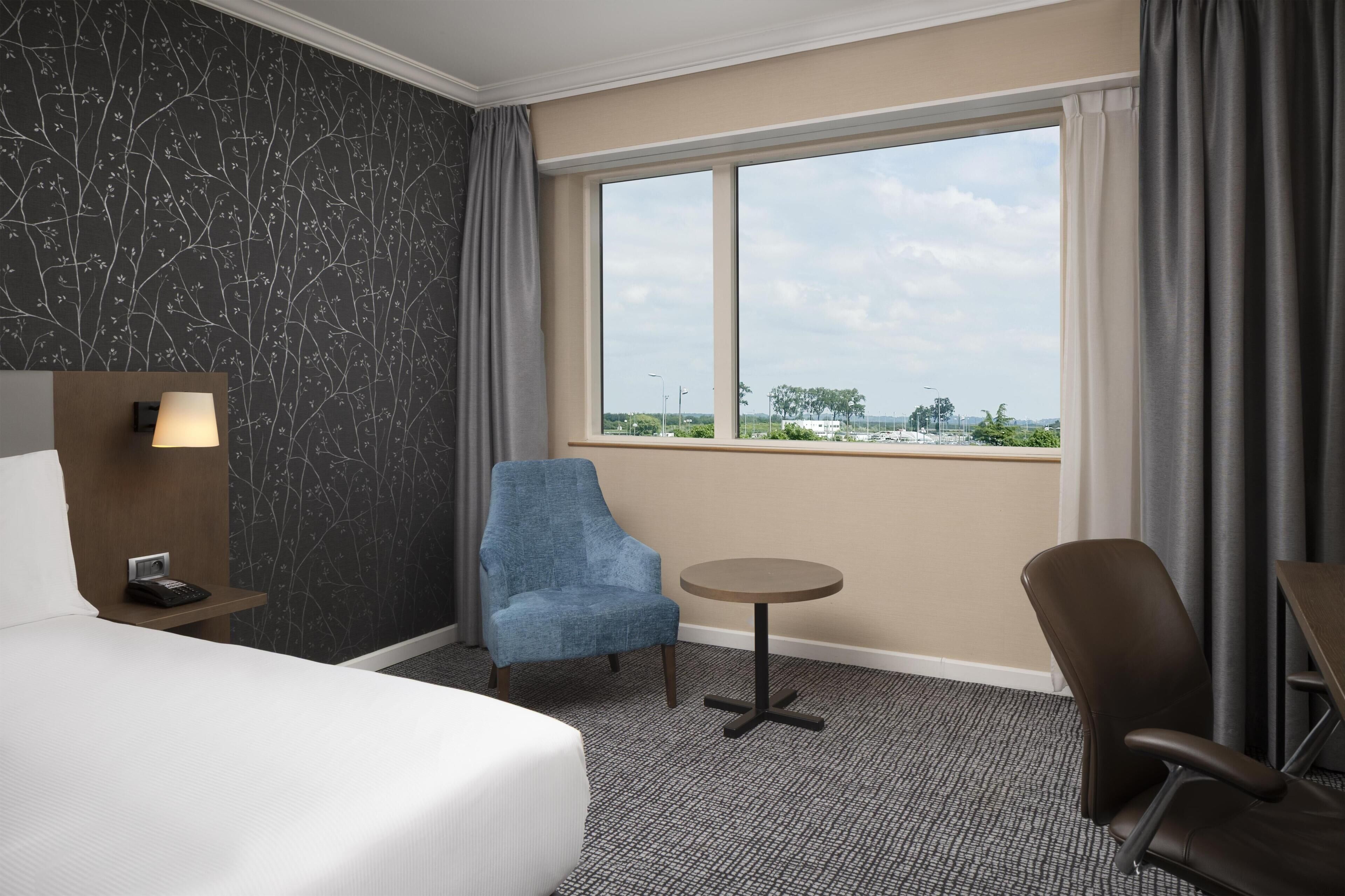 Quarto de hotel com cama, escrivaninha, cadeira, mesinha e vista da cidade através de uma grande janela.
