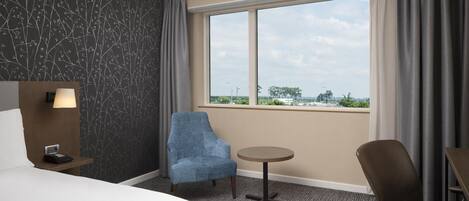 Quarto de hotel com cama, escrivaninha, cadeira, mesinha e vista da cidade através de uma grande janela.
