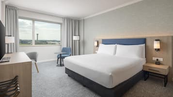 A hotel room with a large bed, a desk, a chair, and a view of the airport.