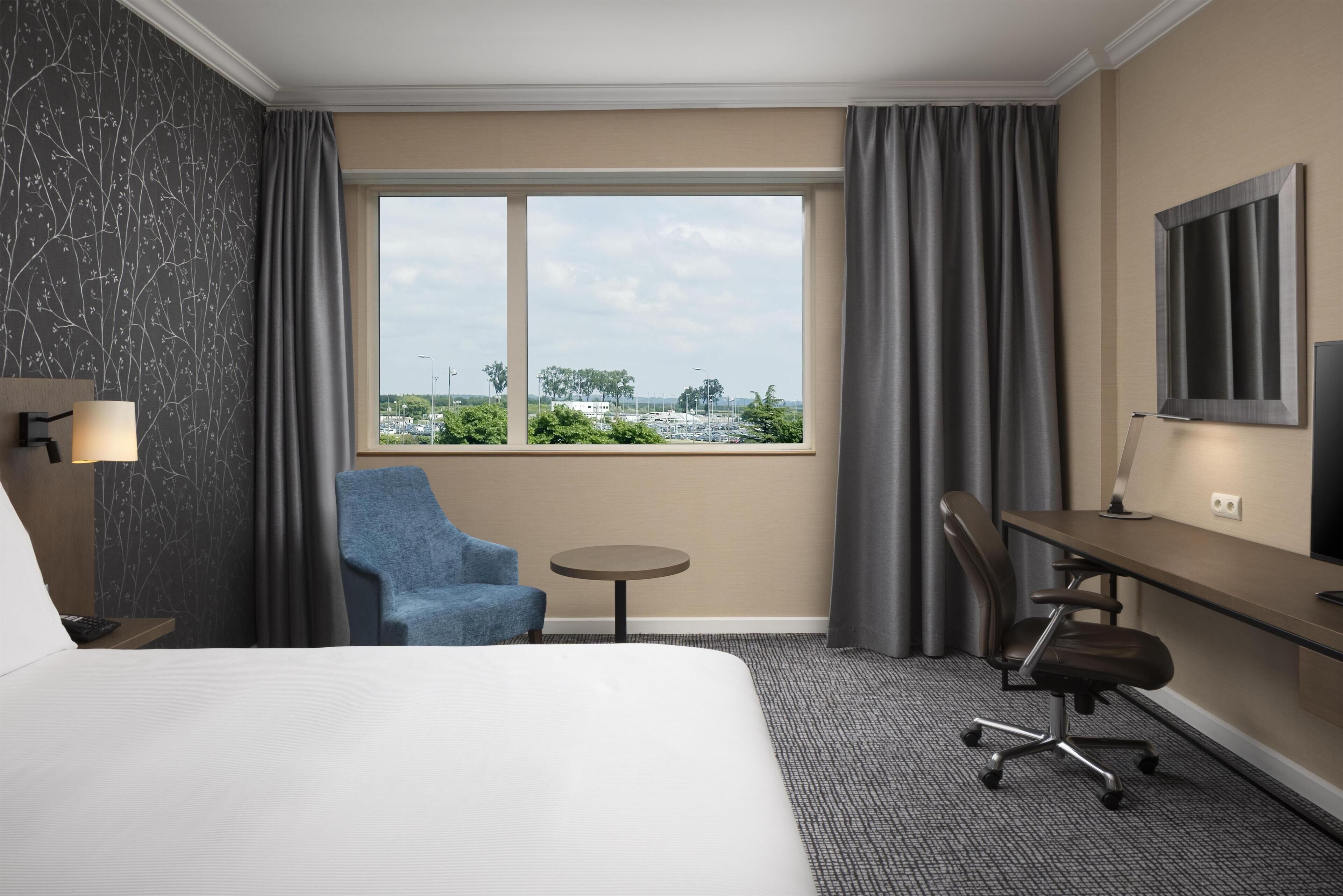 A hotel room with a bed, a desk, a chair, a small round table, and a view of the parking lot.