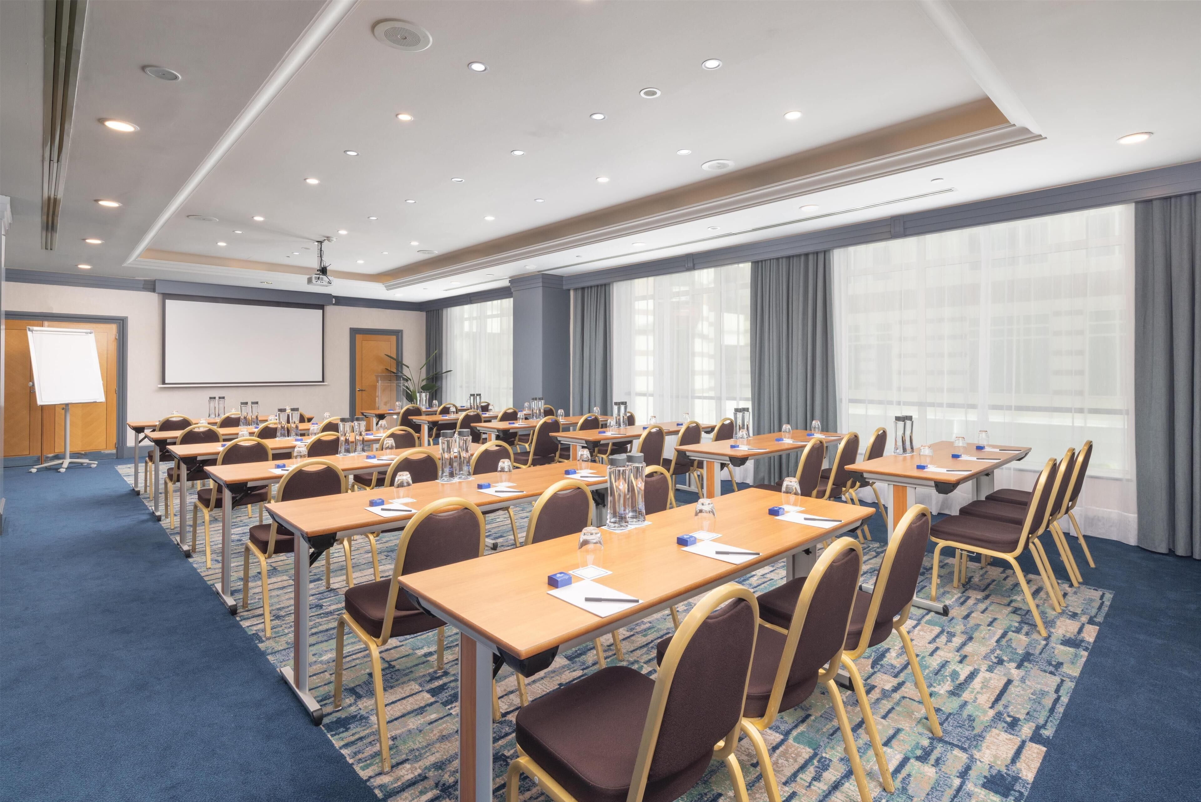 A conference room with multiple tables and chairs, a projector screen, and a large window with curtains.