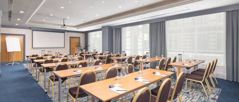 A conference room with multiple tables and chairs, a projector screen, and a large window with curtains.