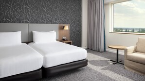 A hotel room with two beds, a nightstand, a lamp, a small table, a sofa, and a large window with a view. - Hilton Paris Charles de Gaulle Airport (Tremblay-en-France)