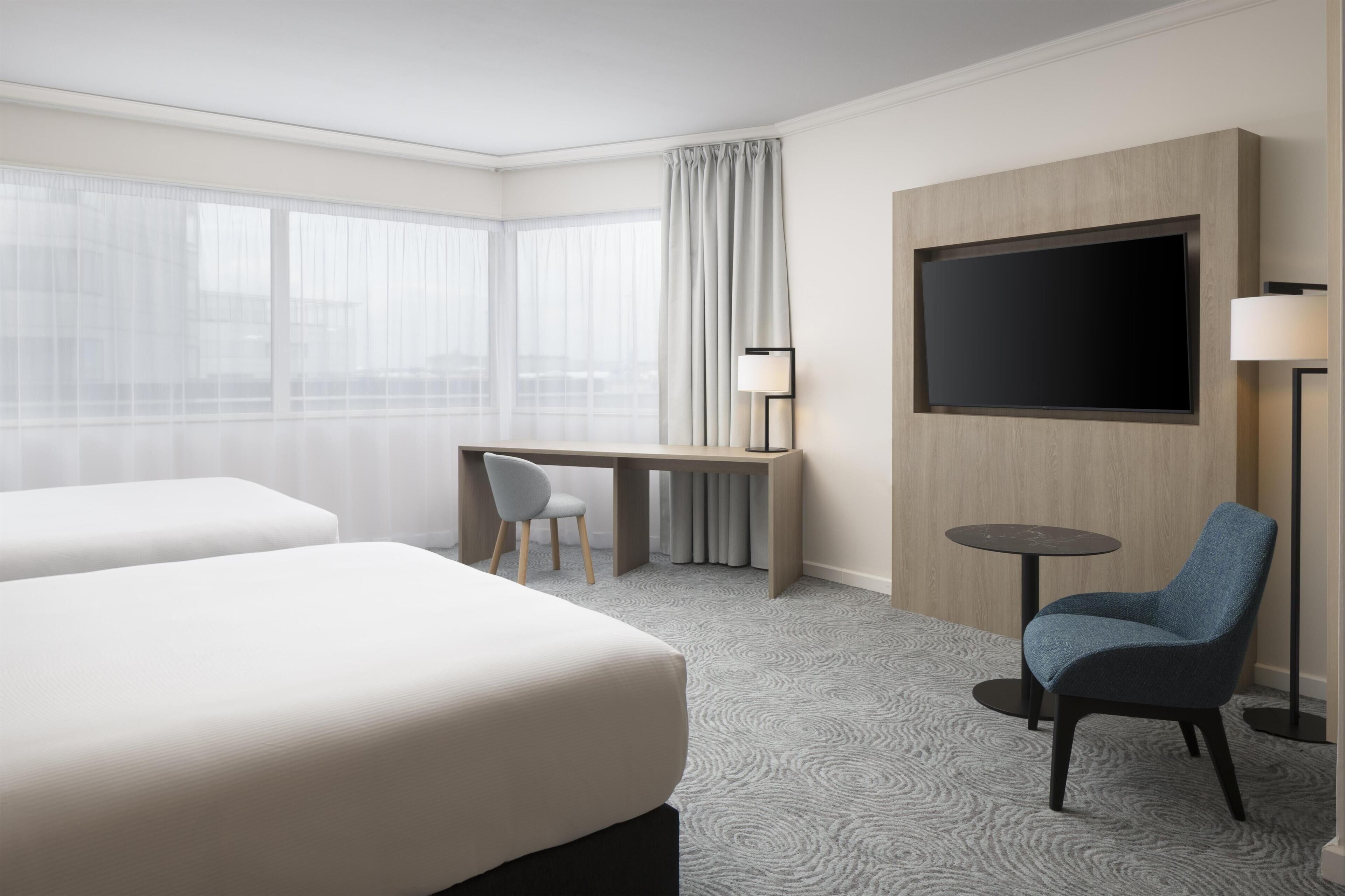 A hotel room with two beds, a desk, a chair, a TV, and a lamp.