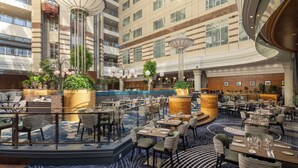 Un amplio vestíbulo de hotel con varias zonas de restauración, grandes plantas en macetas y una barra central.