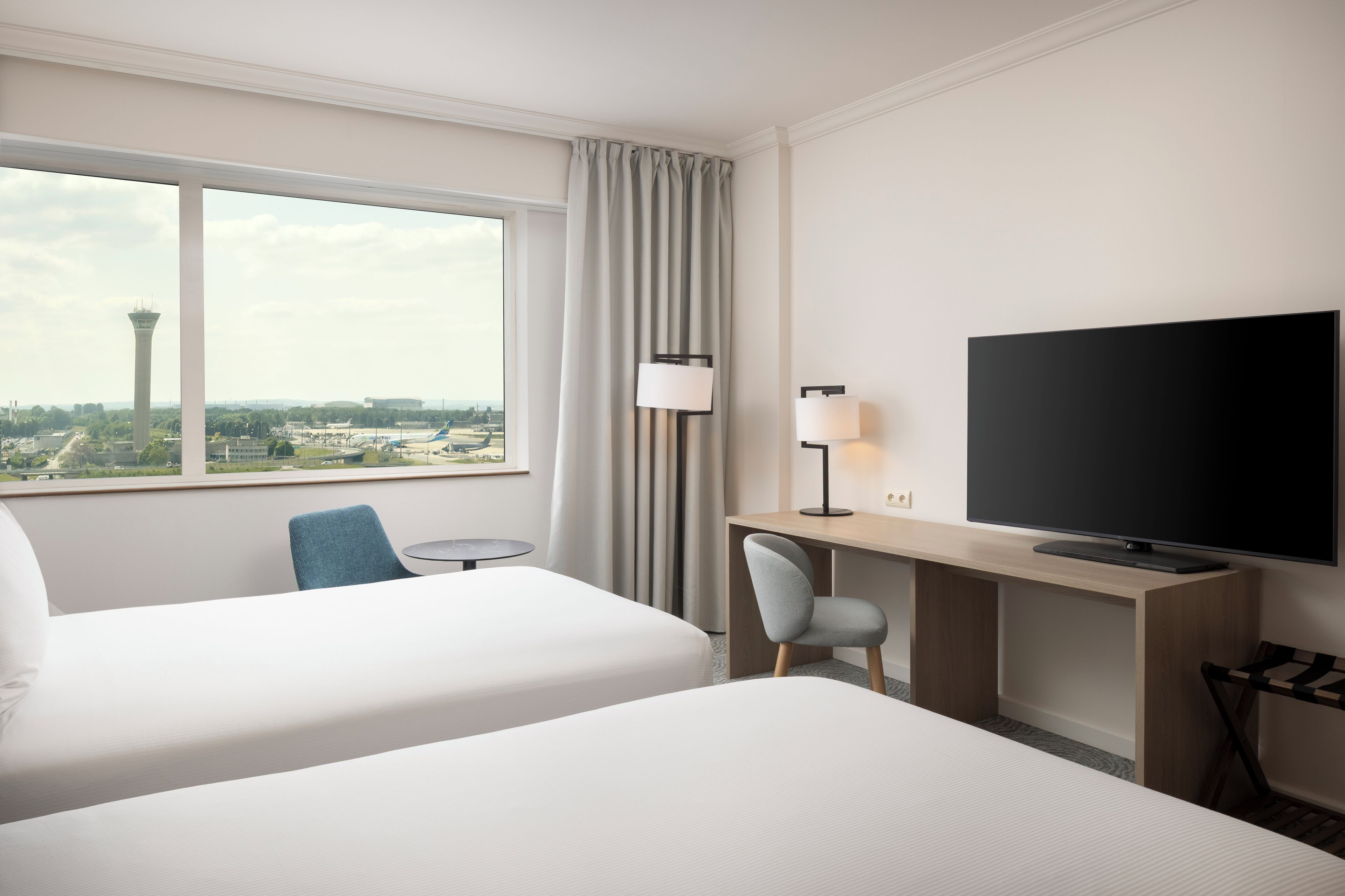 A hotel room with two beds, a desk with a TV, a chair, and a view of a tower and cityscape.
