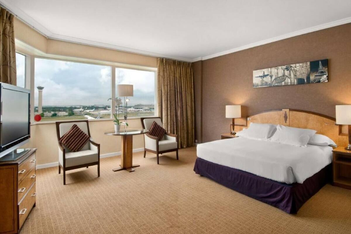 A hotel room with a large bed, two chairs, a TV, and a window with a view.