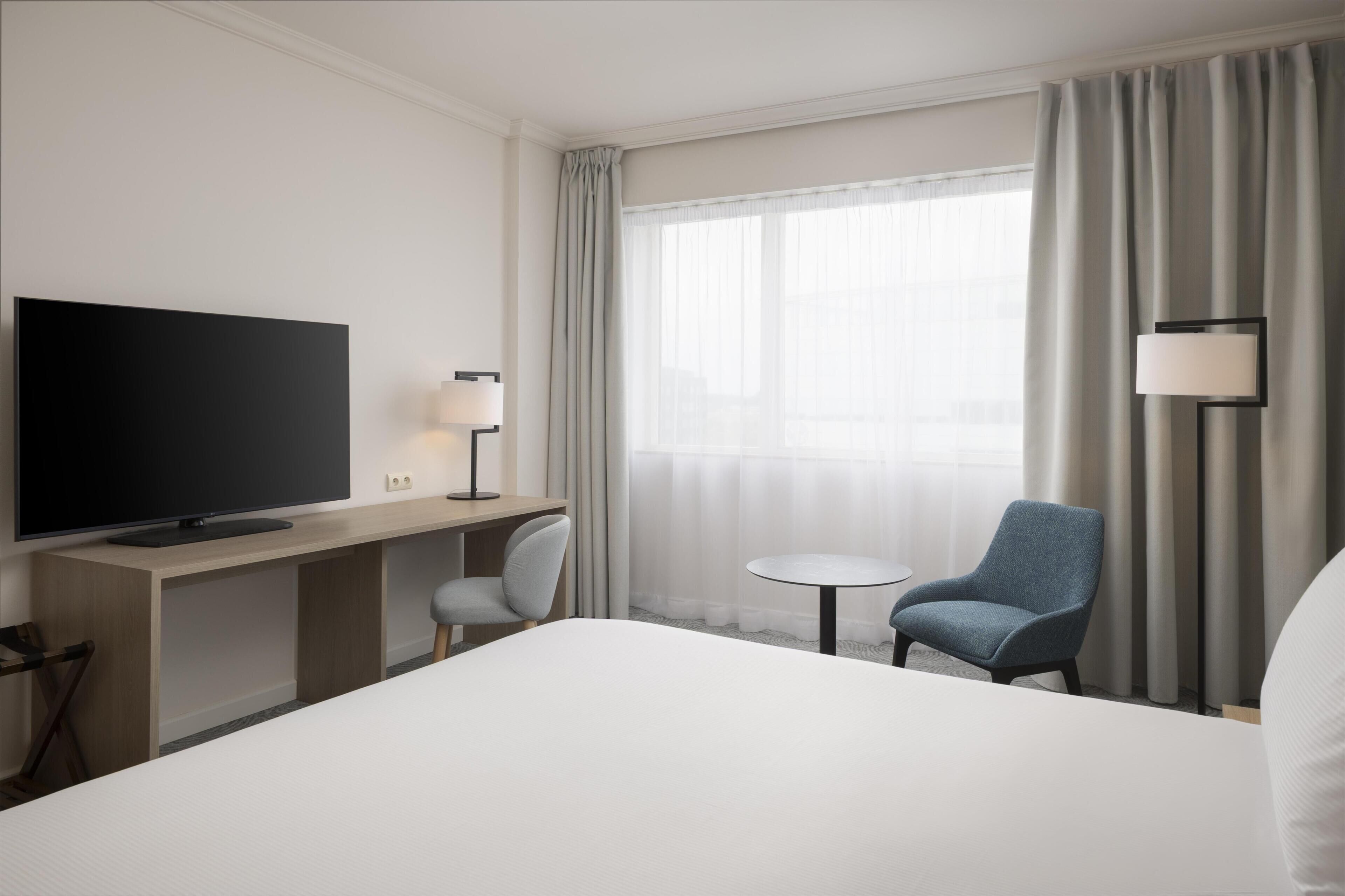 Une chambre d’hôtel équipée d’un téléviseur à écran plat, d’un bureau avec une lampe, d’une chaise, d’une table ronde et d’un lit.