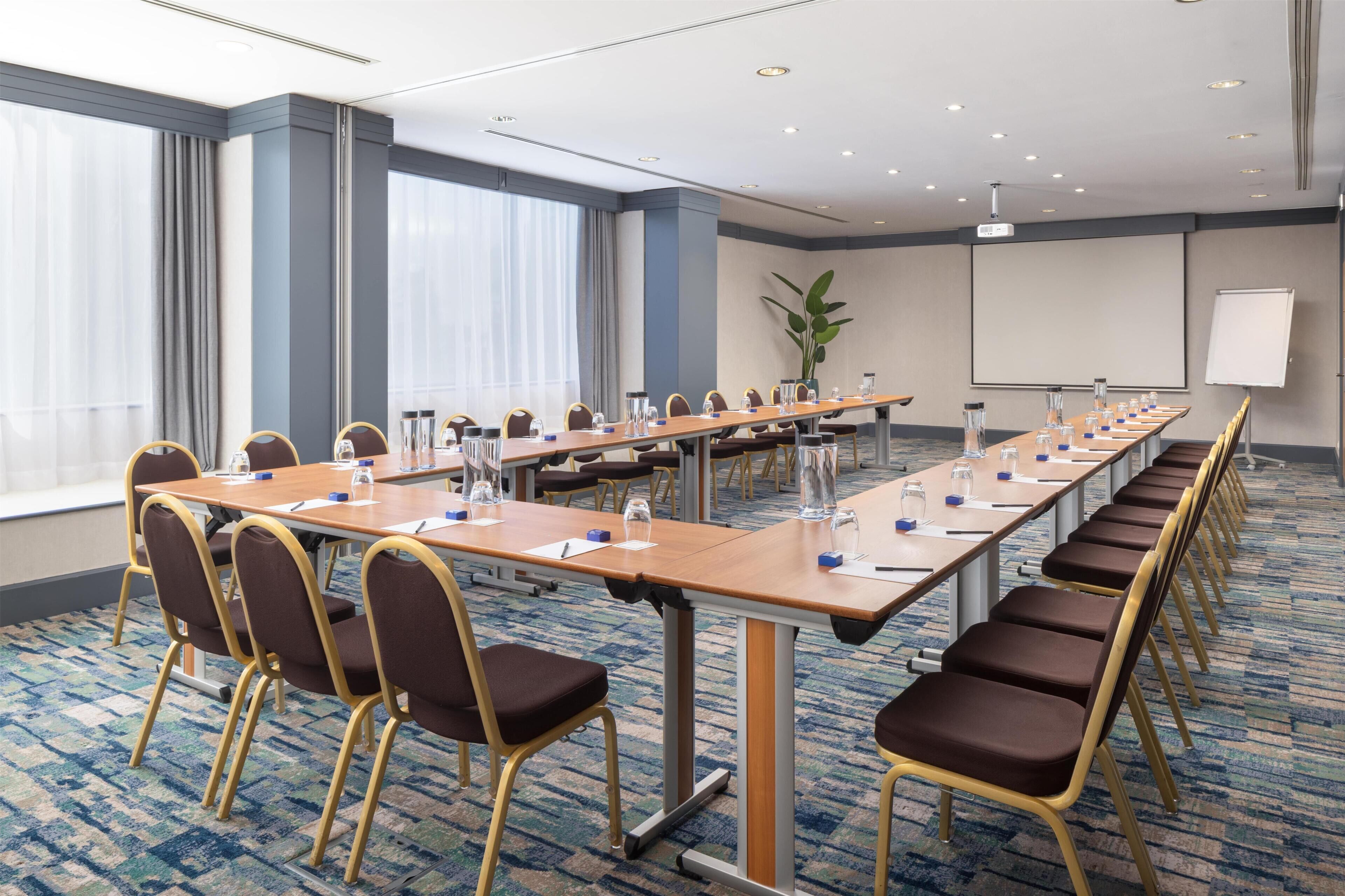 Un salón de conferencias con largas mesas, sillas y una pantalla de proyector.