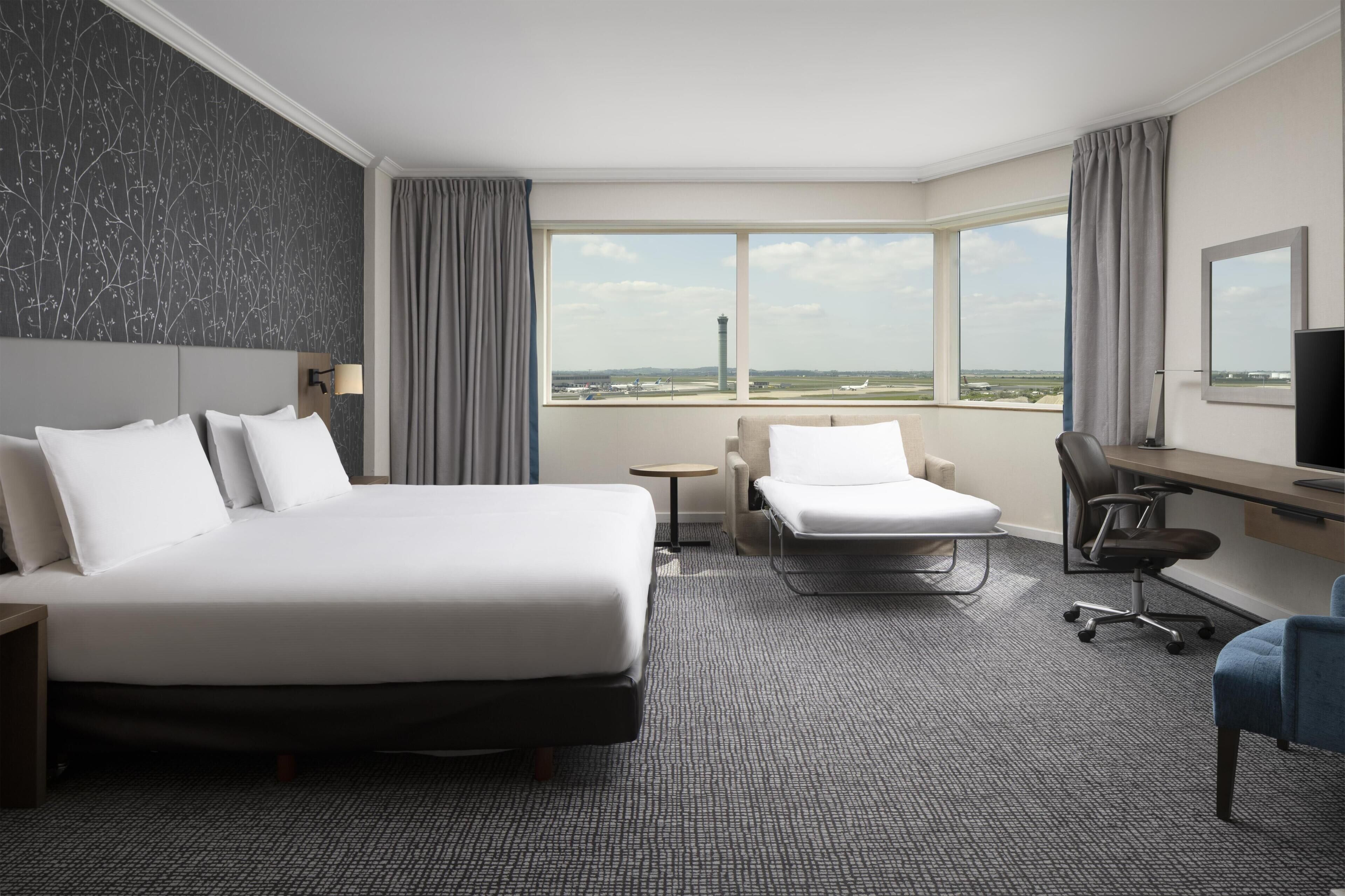 Quarto de hotel com uma cama grande, uma escrivaninha, uma cadeira e vista para o aeroporto.