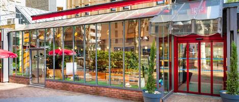 Um restaurante com fachada de vidro, toldo vermelho, mesas ao ar livre e plantas em vasos.