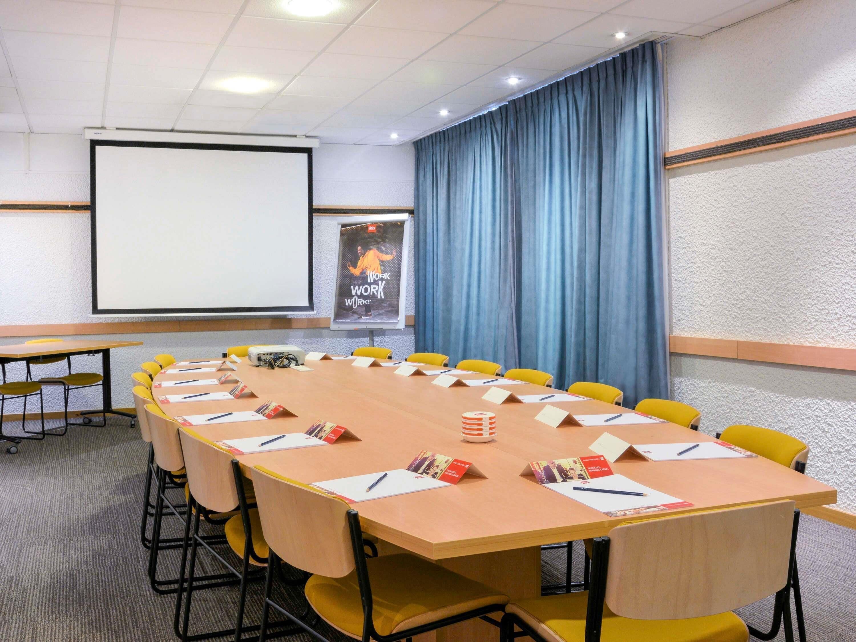 A conference room with a long table, yellow chairs, name cards, a projector screen, and a poster on the wall.