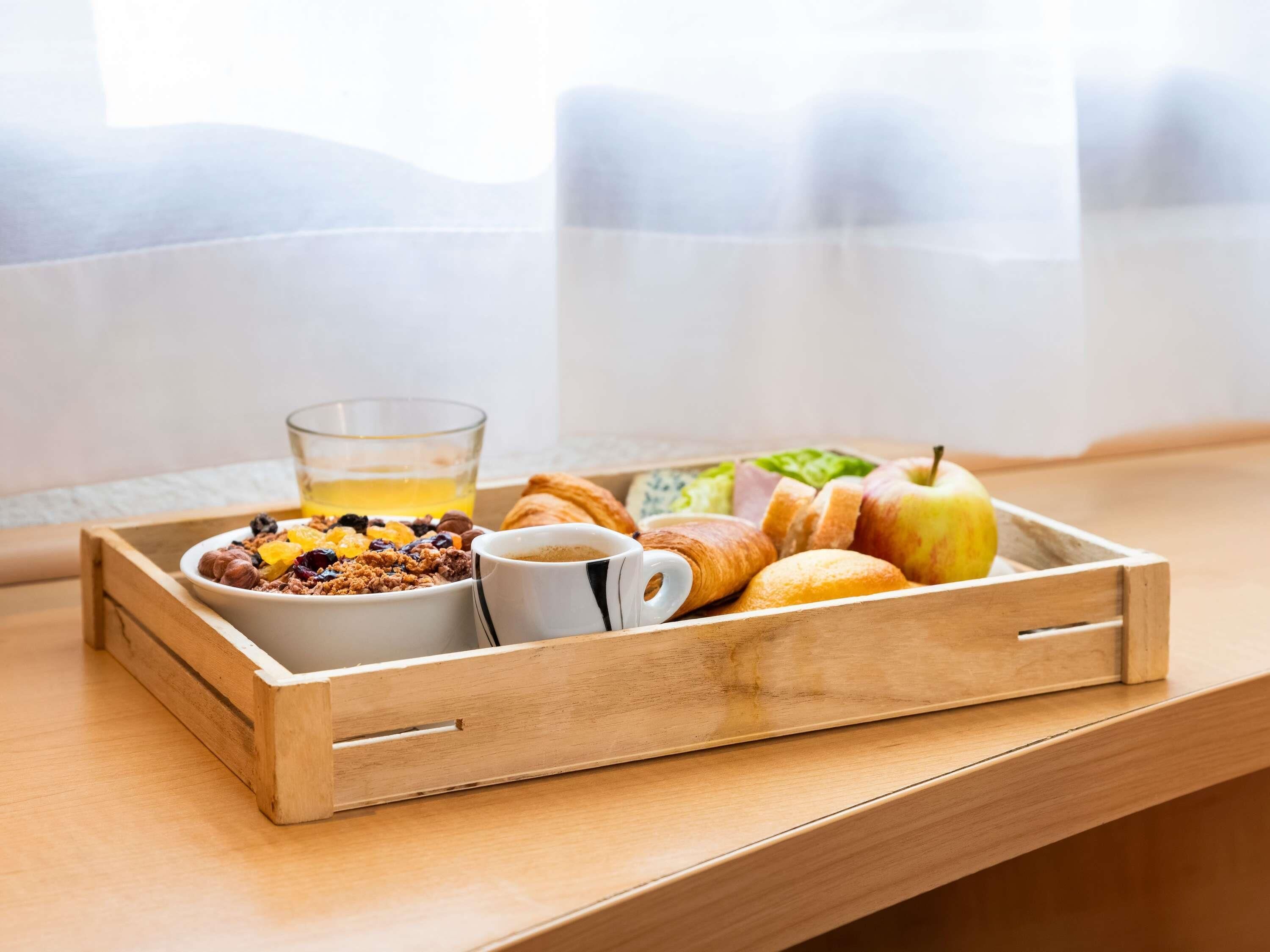 A wooden tray with breakfast items including cereal, a glass of orange juice, a croissant, an apple, and a cup of coffee on a wooden table near a window.