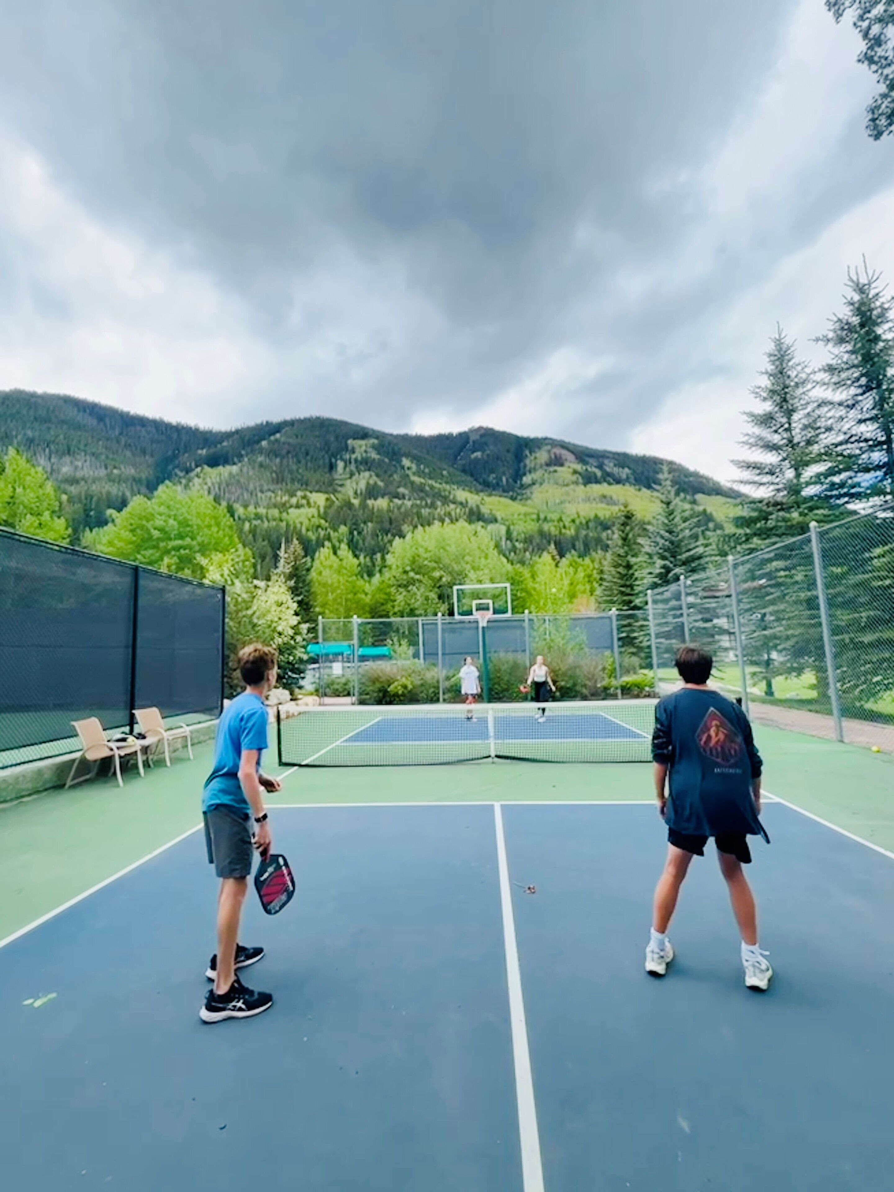 An outdoor tennis court with a net, surrounded by chairs and a fence, with mountains and trees in the background.