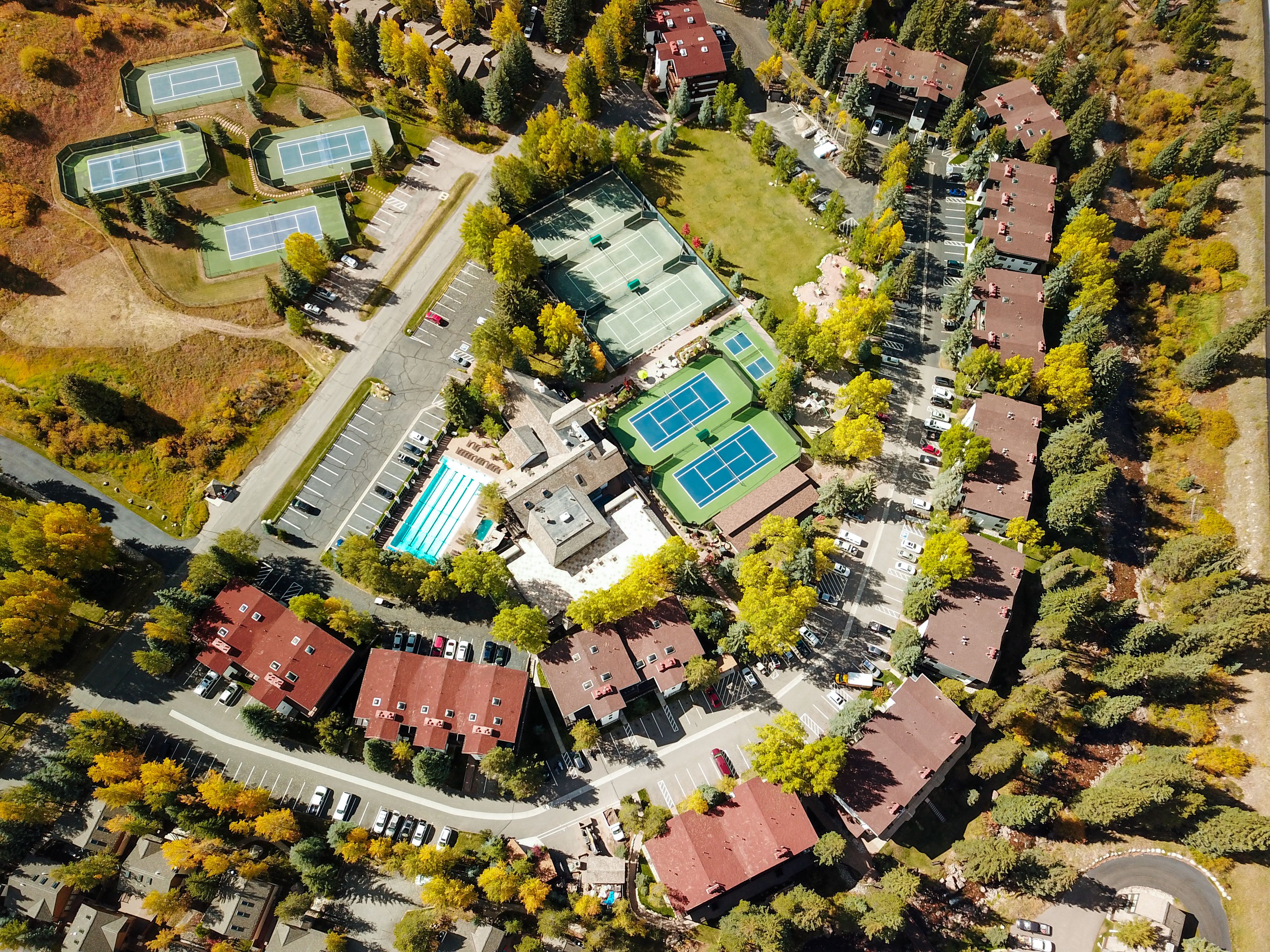 An aerial view of a residential area with multiple houses, a swimming pool, tennis courts, and a parking lot.