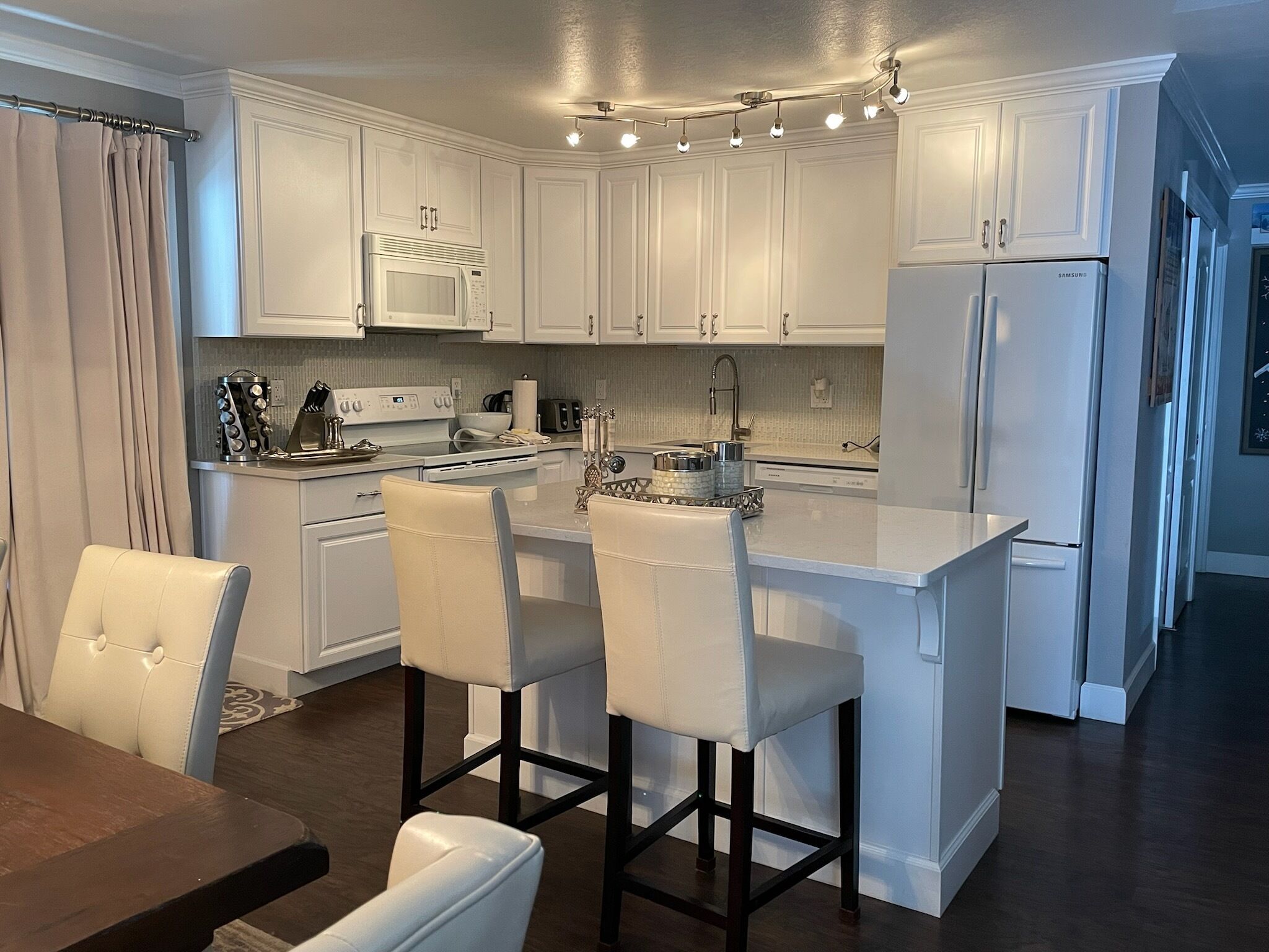 Cocina moderna con armarios blancos, una isla central con taburetes altos, electrodomésticos de acero inoxidable e iluminación colgante.