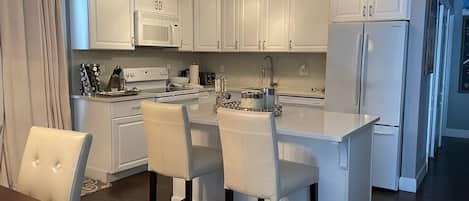 Cocina moderna con armarios blancos, una isla central con taburetes altos, electrodomésticos de acero inoxidable e iluminación colgante.