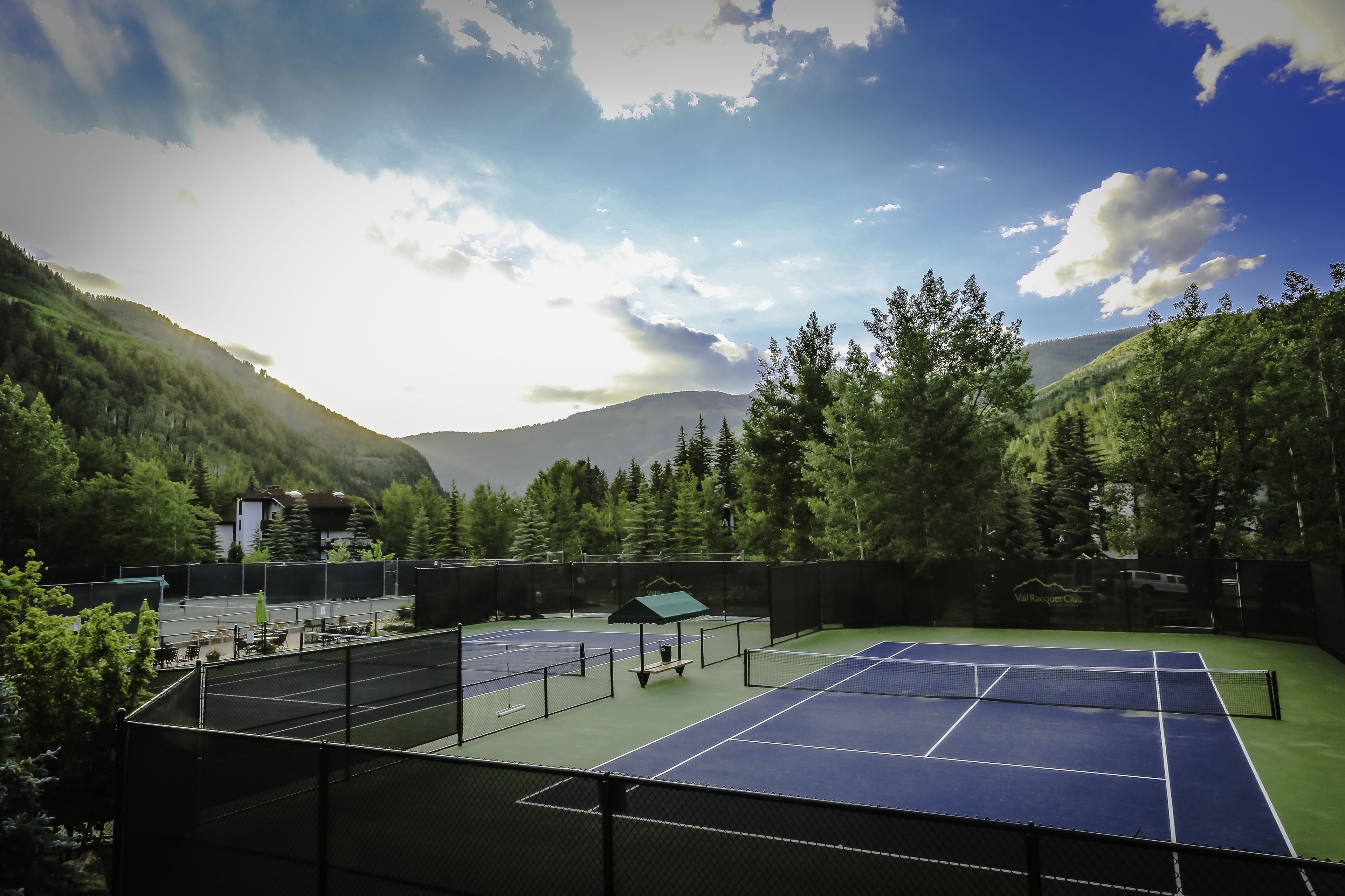 Cancha de tenis al aire libre con red, rodeada de árboles y una montaña al fondo.