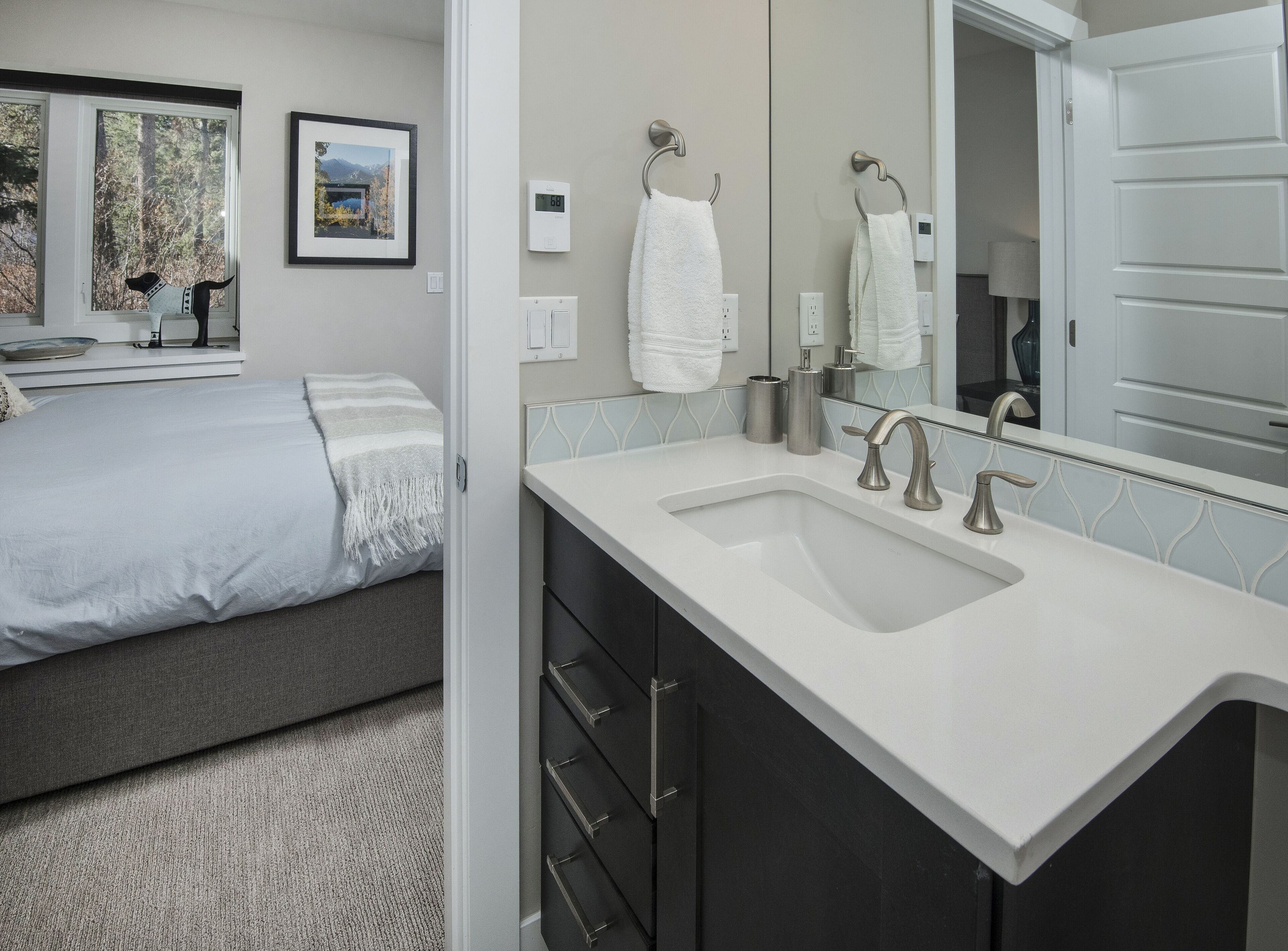 Un baño moderno con un lavamanos grande, un espejo y un inodoro. Hay una cama con cabecera gris y vistas a árboles a través de la ventana.